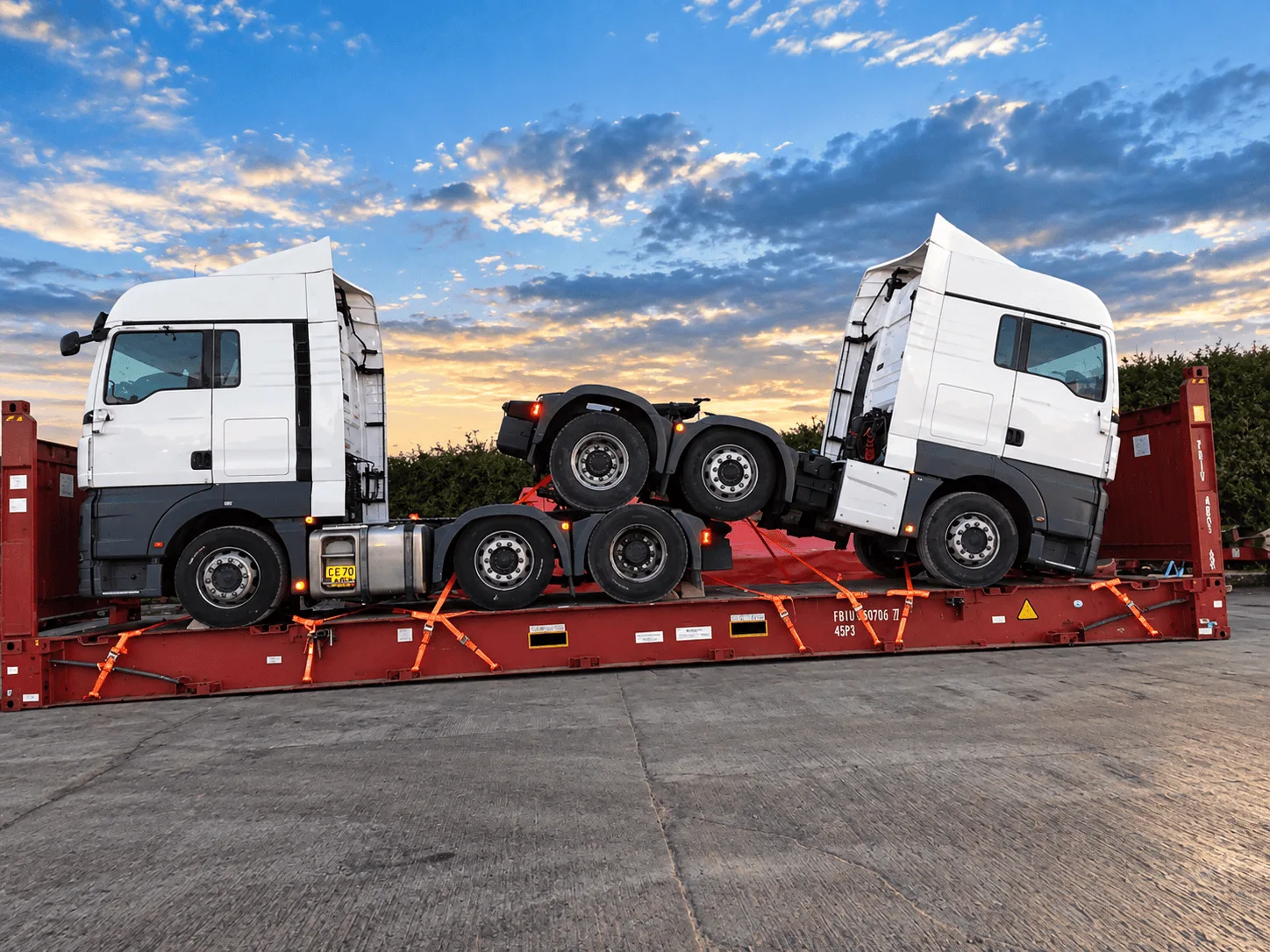 Two white prime mover trucks securely transported on a red flat-rack platform for international logistics.